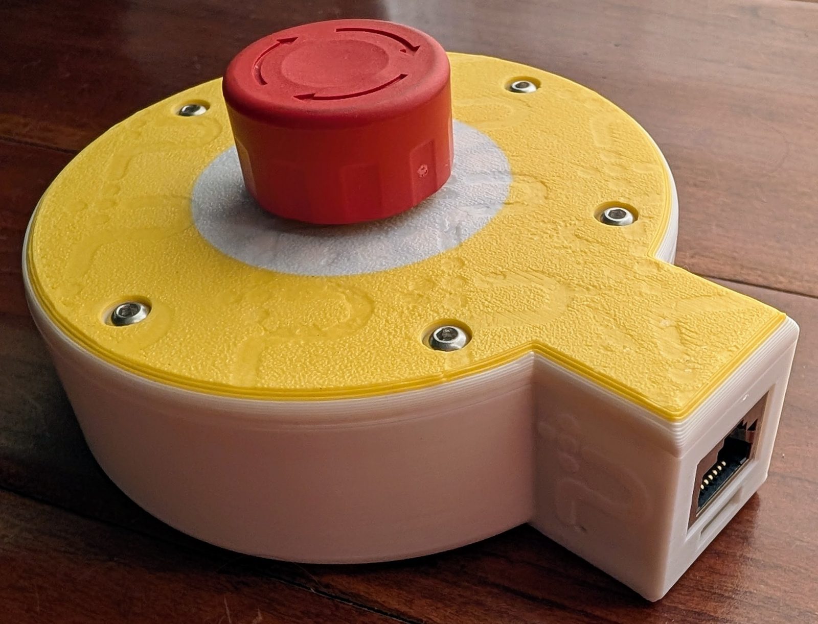 The Protective Stop remote: a red mushroom stop button on a 3D-printed yellow and white enclosure, with an ethernet socket in the side.